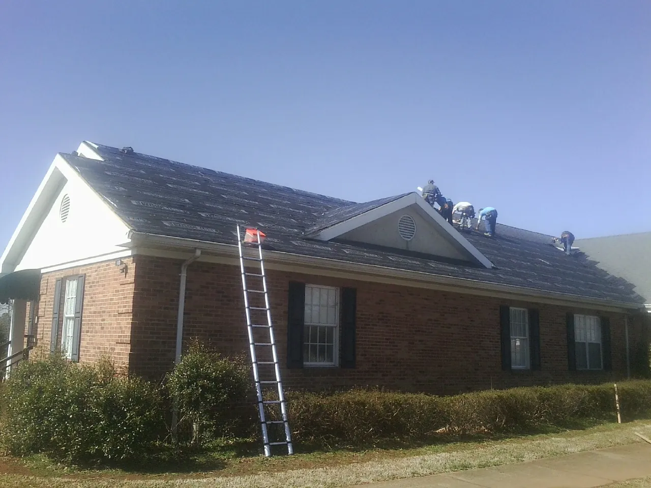 Crew tearing shingles off a brick ranch roof with a ladder set — Metal Roof Repair work by our crew in Iron City