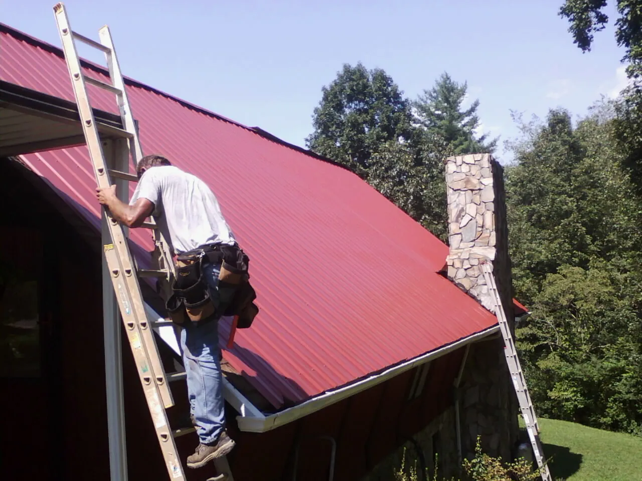Roofer on a ladder beside a red metal roof and stone chimney — Soffit & Fascia Repair by Roofing Brothers in Iron City
